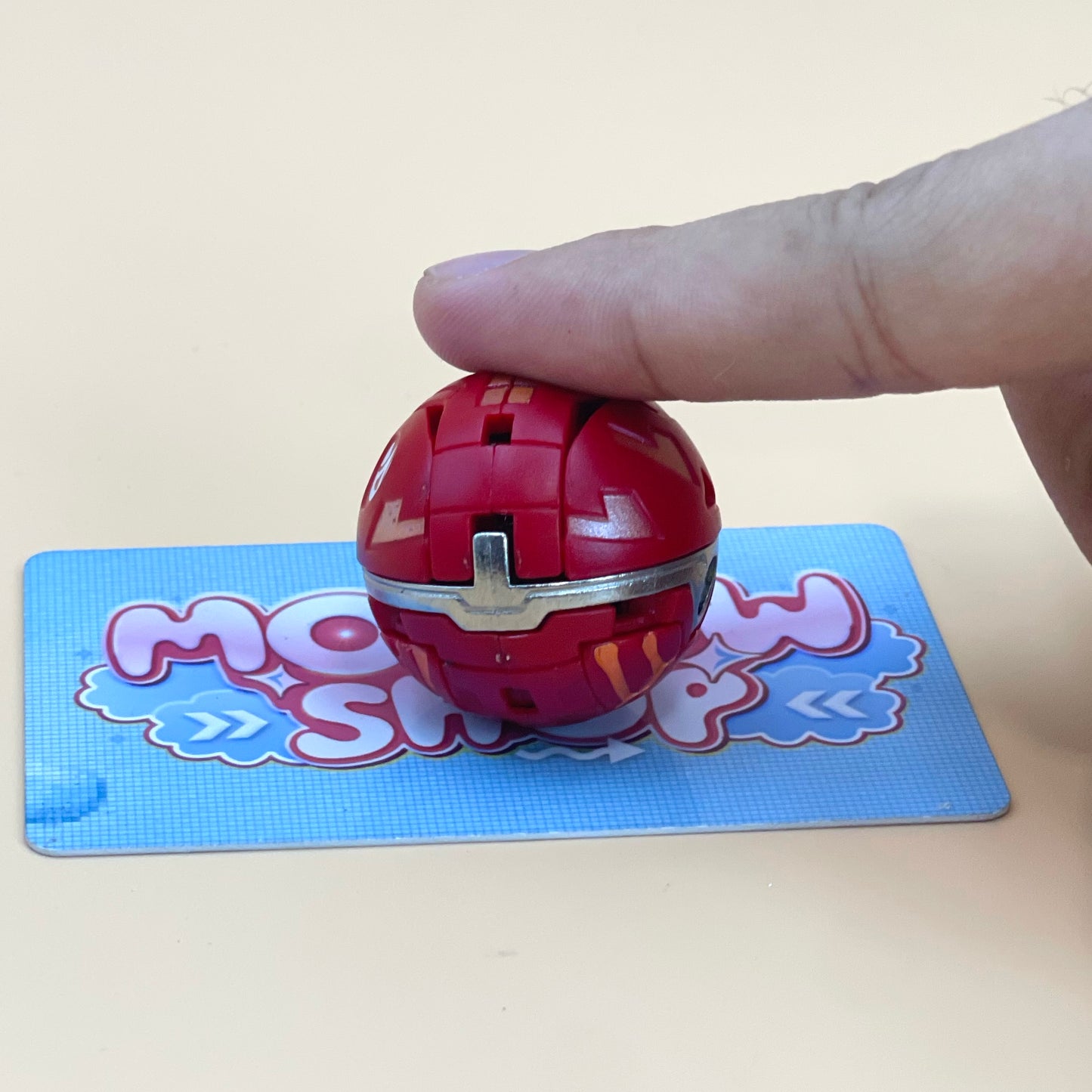 A hand is holding a red ball with a metallic surface, which is resting on a blue card with a cartoon character design.