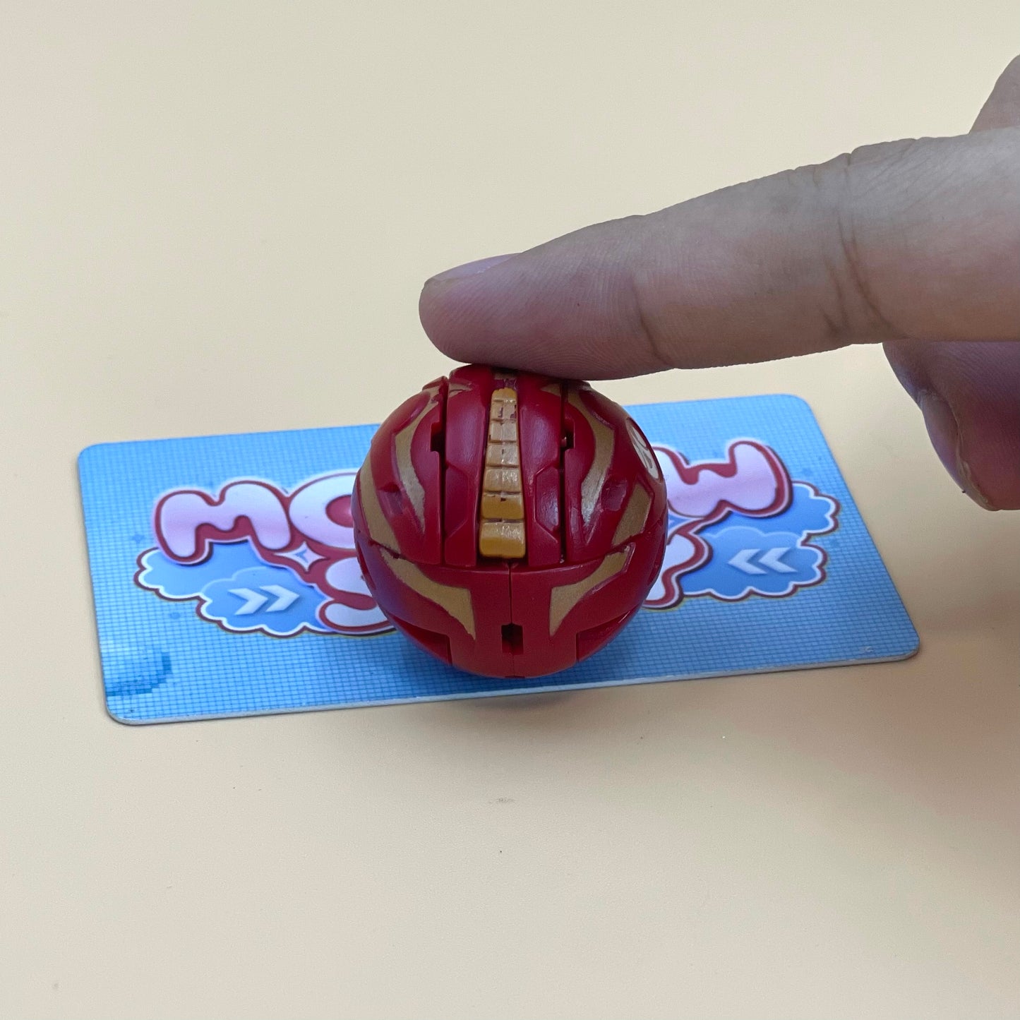 A person's hand is holding a red and gold toy, which appears to be a toy car, on a blue and white card with a cartoon character design.