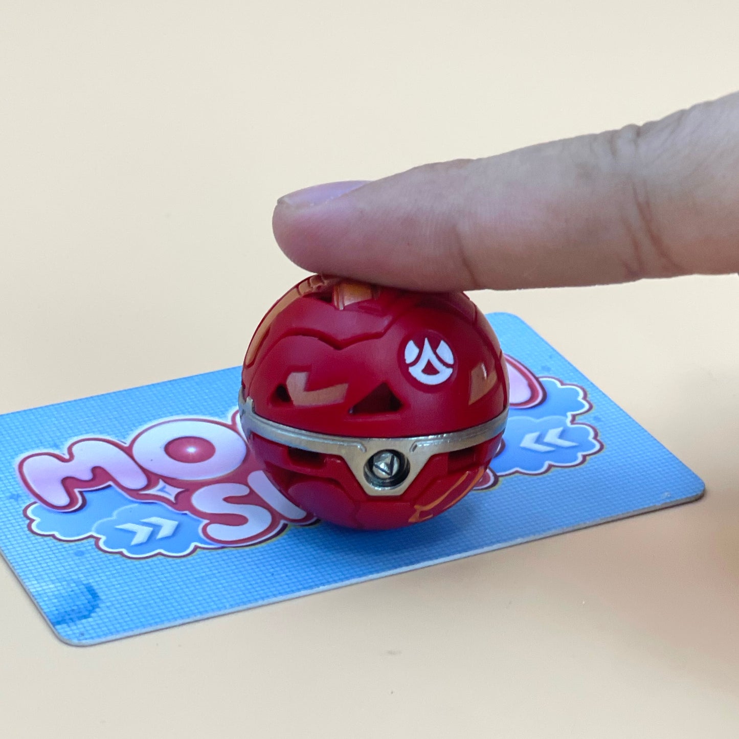 A hand is holding a red ball with a silver clasp, which is placed on a blue card with a cartoon character design.