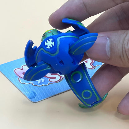 A hand is holding a blue and green toy in the shape of a ball, with a card featuring a cartoon character in the background.