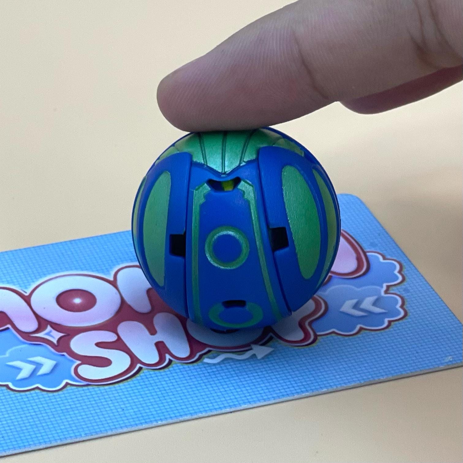 A hand is holding a blue and green toy, which appears to be a ball or a toy ball, on a blue surface with a partially visible text logo.