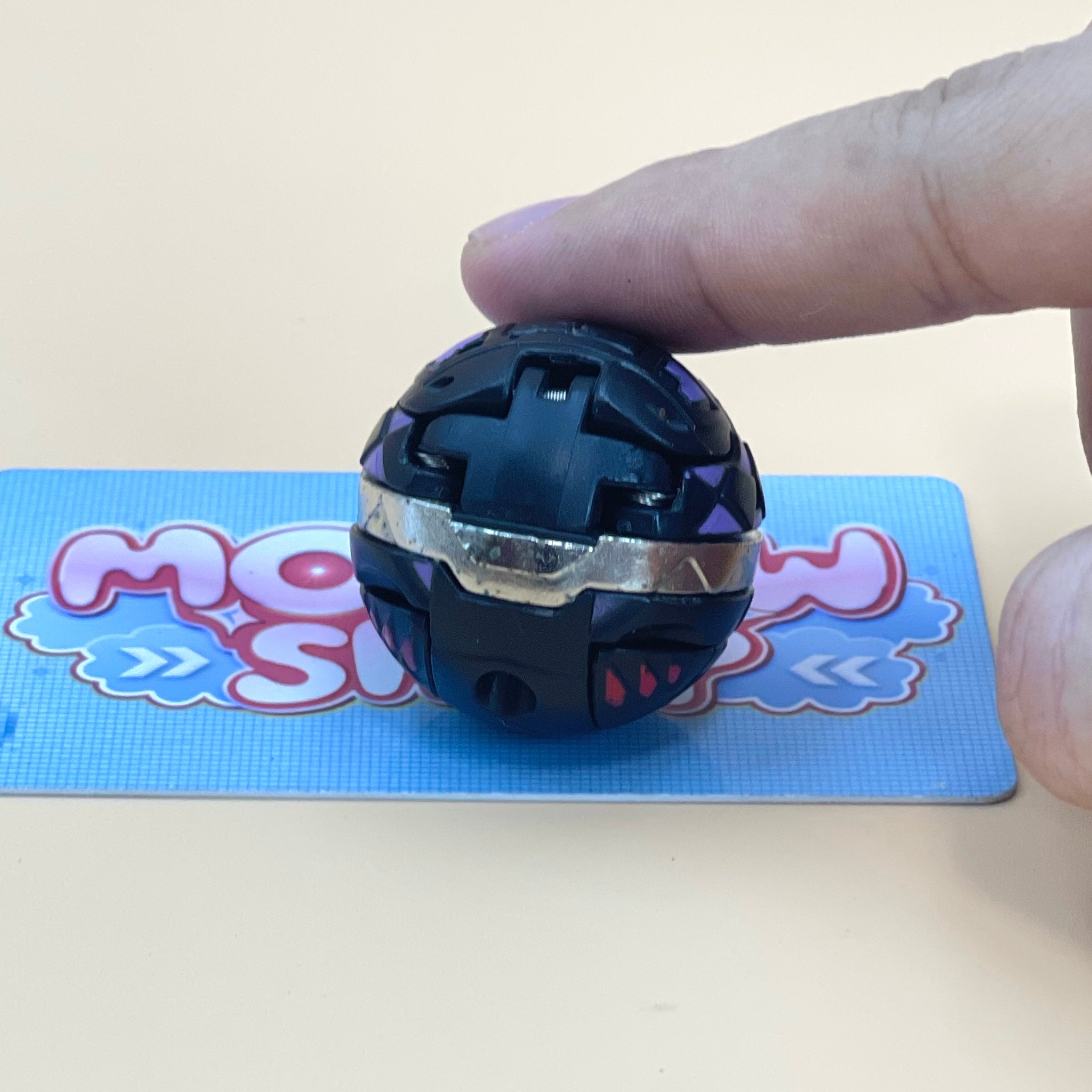 A person's hand is holding a small, black and purple ball with a silver ring on it, placed on a blue surface with a partially visible card or sign in the background.