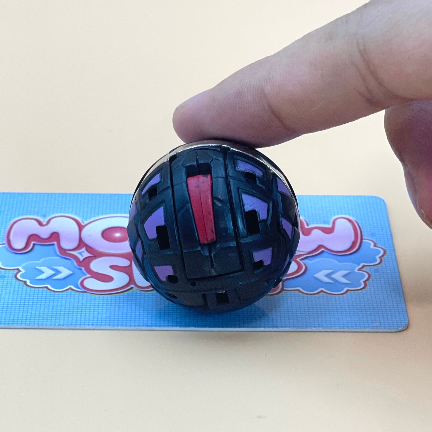 A hand is holding a black and purple toy, which appears to be a ball or a small toy, on a blue surface with a partially visible text logo.