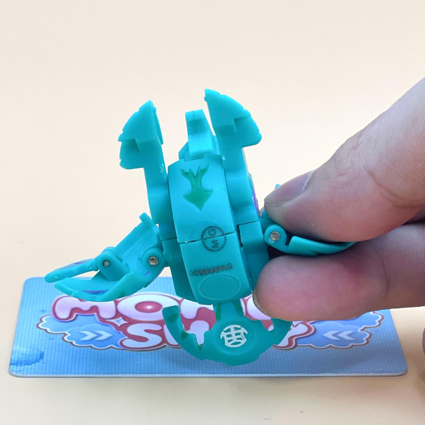 A hand is holding a small, light blue toy with a green design on it, which appears to be a toy car or toy vehicle.
