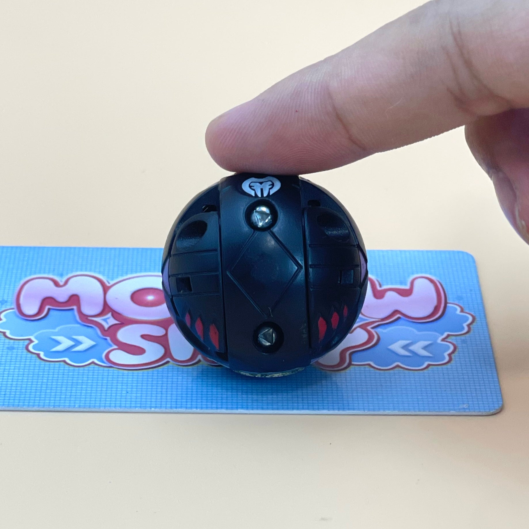 A person's finger is holding a black, round toy with a white light bulb on it, placed on a blue and pink card with a cartoon character design.