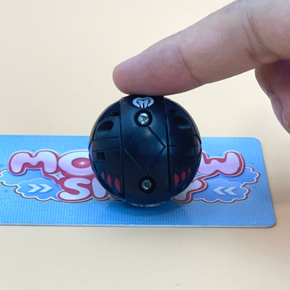 A person's finger is holding a black, round toy with a white light bulb on it, placed on a blue and pink card with a cartoon character design.
