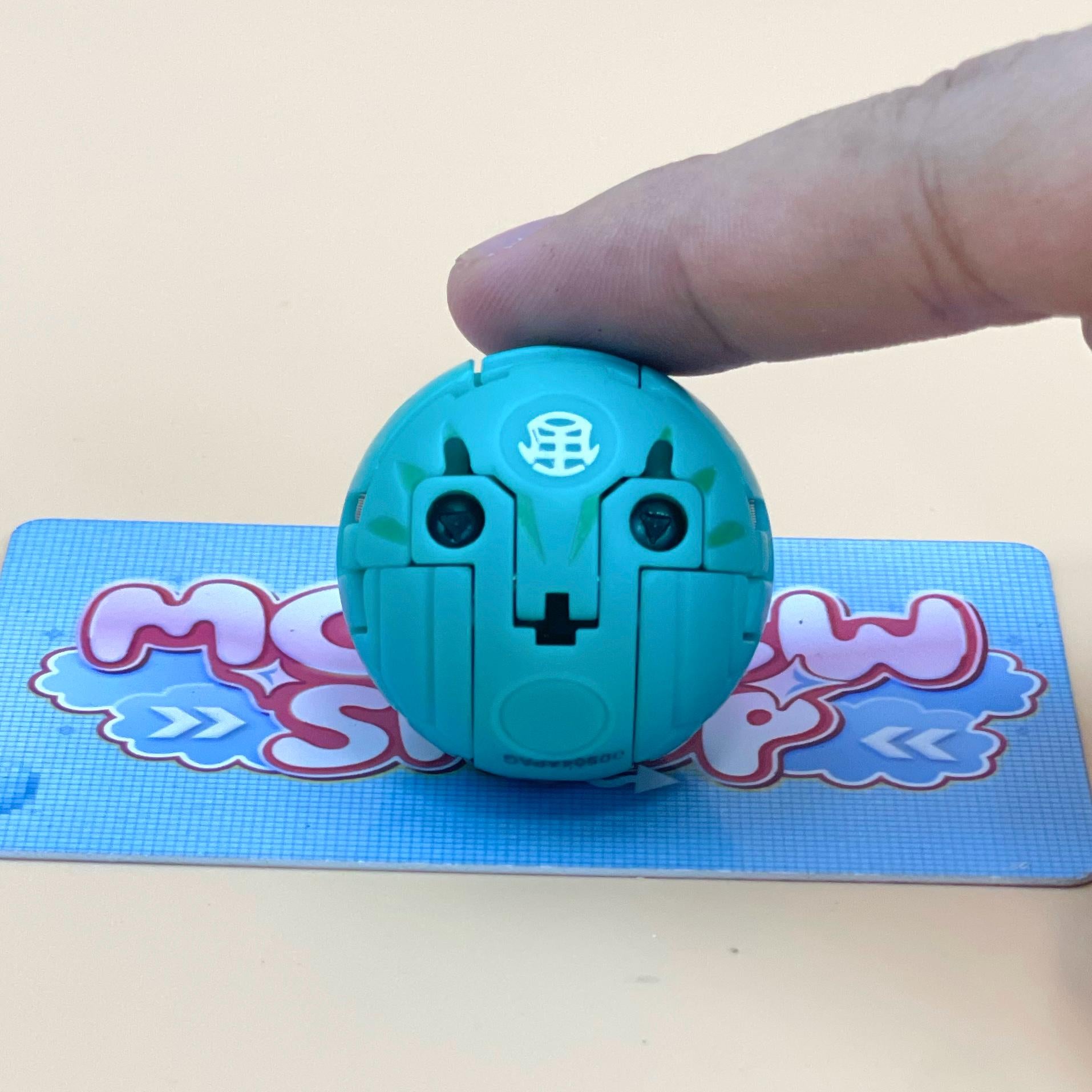 A hand is holding a small, light blue toy with a face design on it, which is placed on a blue and white checkered surface.